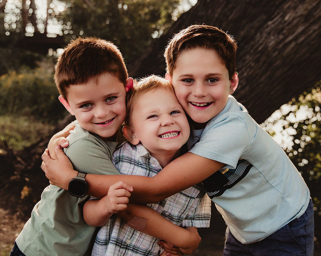 Sioux Falls family photography of three brothers hugging at sunset by a river, older boys wrapping the youngest in a playful embrace while all smile mischievously at the camera