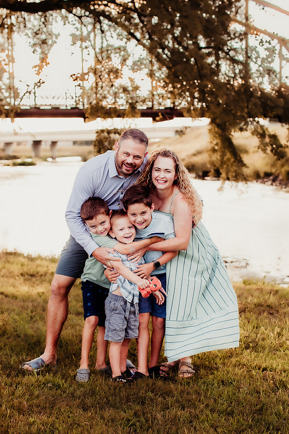 Sioux Falls family of five huddled together on the Big Sioux River shoreline, smiling and cuddling closely in a playful group hug during a lifestyle photography session