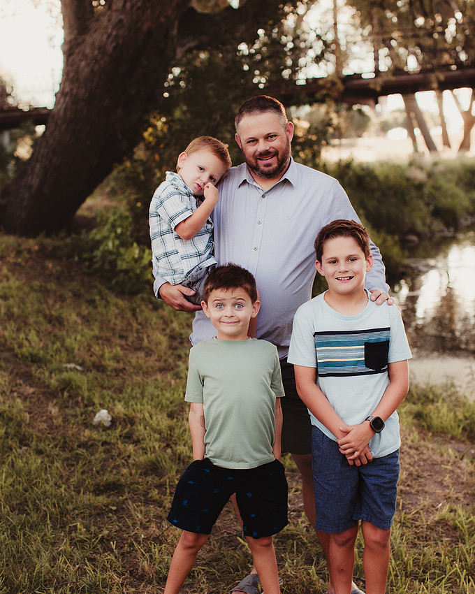 Sioux Falls family photo of a father with his three sons by a river at sunset, youngest child in his arms while two older boys stand in front with playful expressions, bridge and trees in background