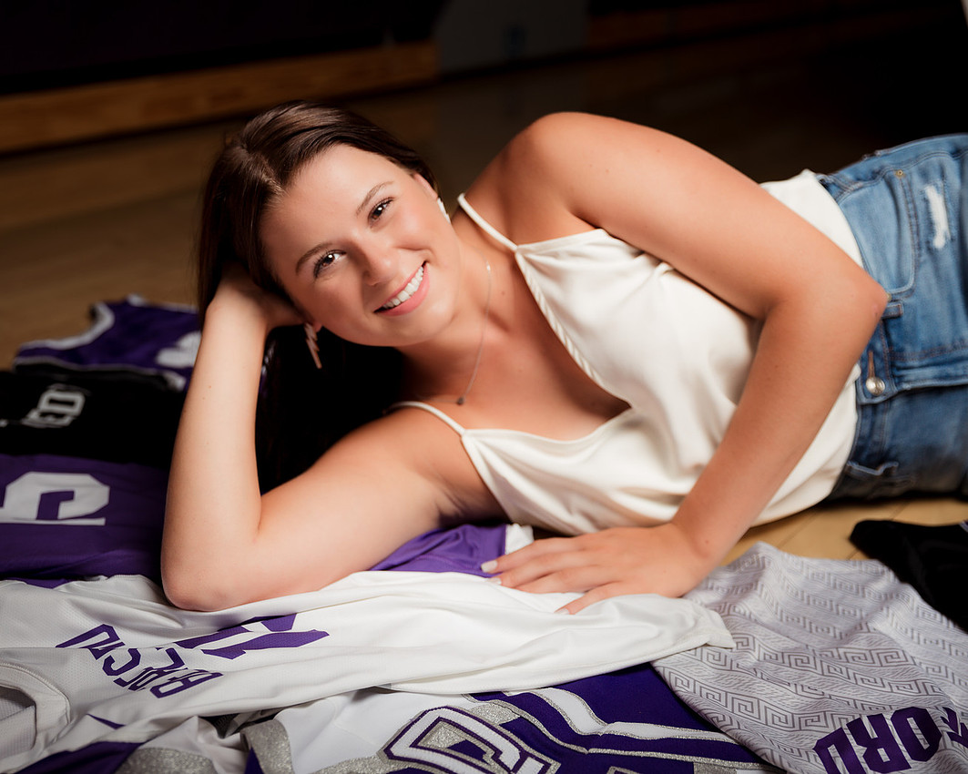 High school senior girl lying across multiple sports jerseys from volleyball, basketball, and track, wearing a dressy shirt and jeans with soft glam lighting highlighting her strength and personality