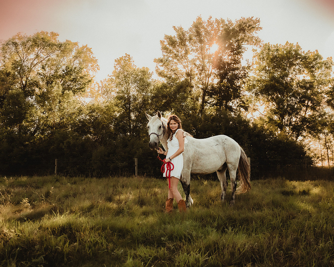 High school senior girl in a short cotton dress and cowboy boots leading a bridled horse through a grassy pasture in South Dakota