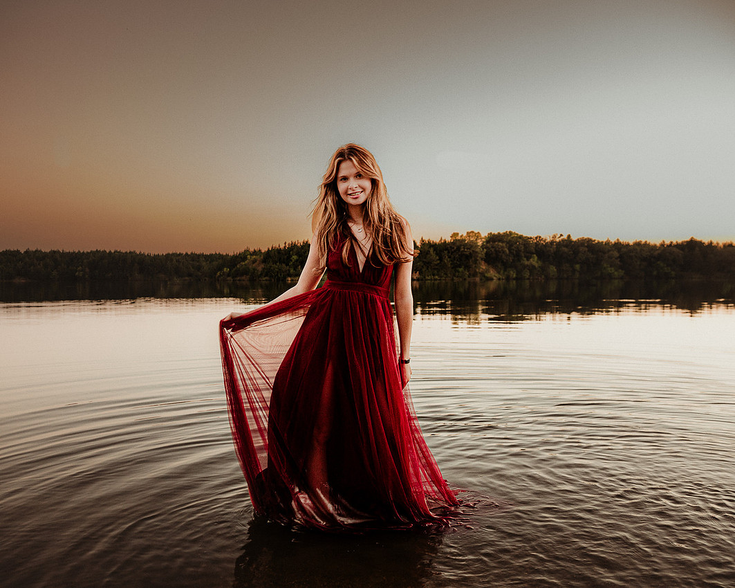 Sioux Falls senior photographer capturing high school senior girl in long red dress standing in lake at sunset with dramatic lighting
