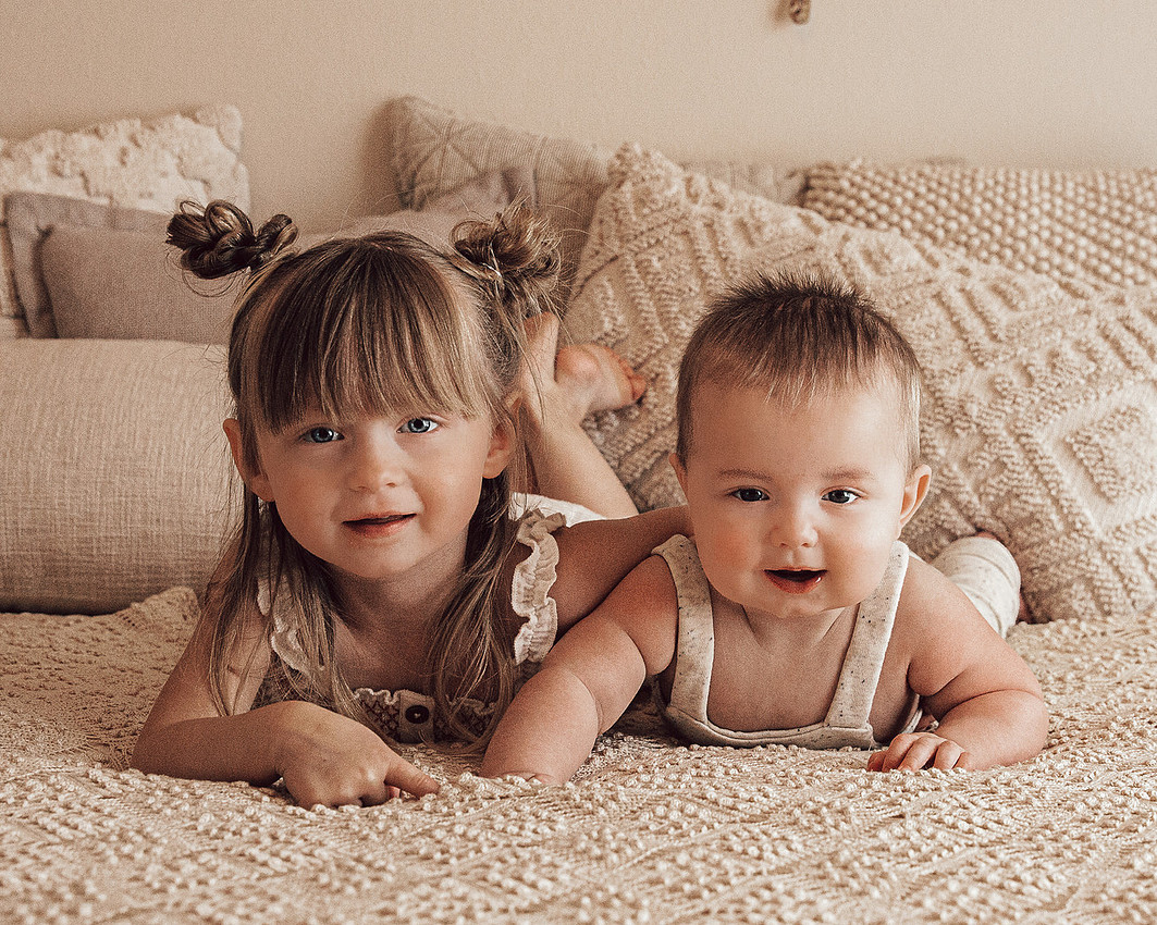 baby and toddler sibling milestone portrait lying on neutral bed during lifestyle children photography session near Sioux Falls South Dakota
