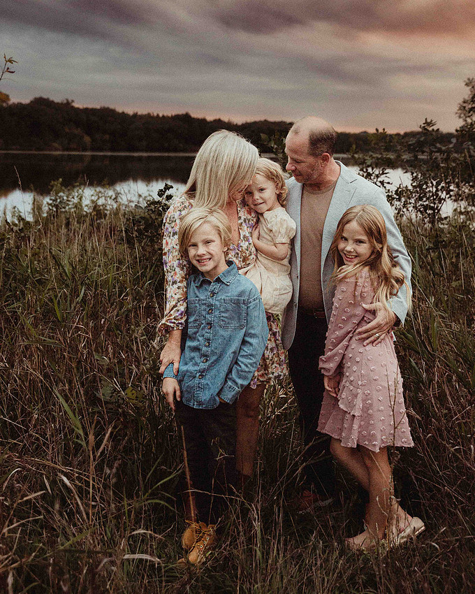 Parents admiring their youngest child in a grassy field by the lake, all part of a family of five.