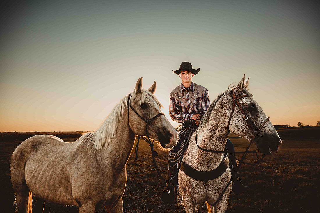 High school senior boy sitting on a saddled horse while holding another horse on a lead rope, backlit by a golden South Dakota sunset