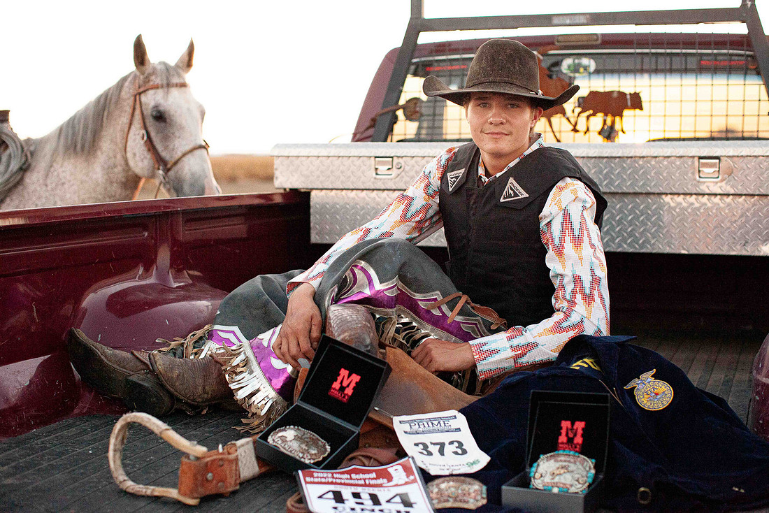 High school senior boy sitting in the back of a pickup truck surrounded by rodeo gear, wearing cowboy hat and chaps with his horse in the background at sunset