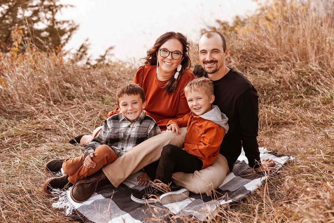 Family of four sitting on a black and white buffalo check blanket during a fall session, wearing coordinated outfits in rust, khaki, black, and green, smiling and laughing together