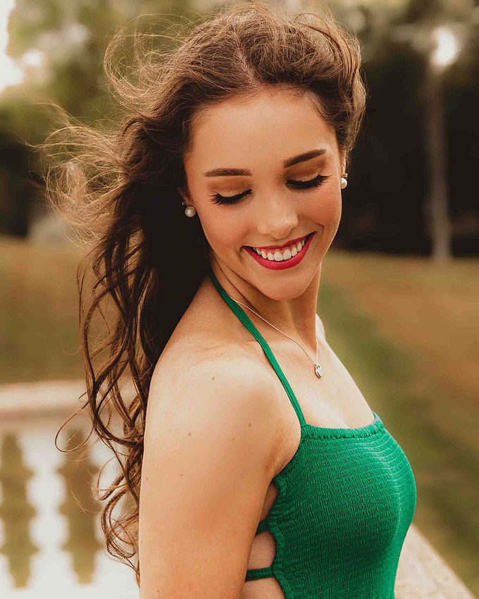 Close-up of a high school senior girl in a green dress looking down and smiling, wearing pearl earrings and red lipstick with soft flowing hair