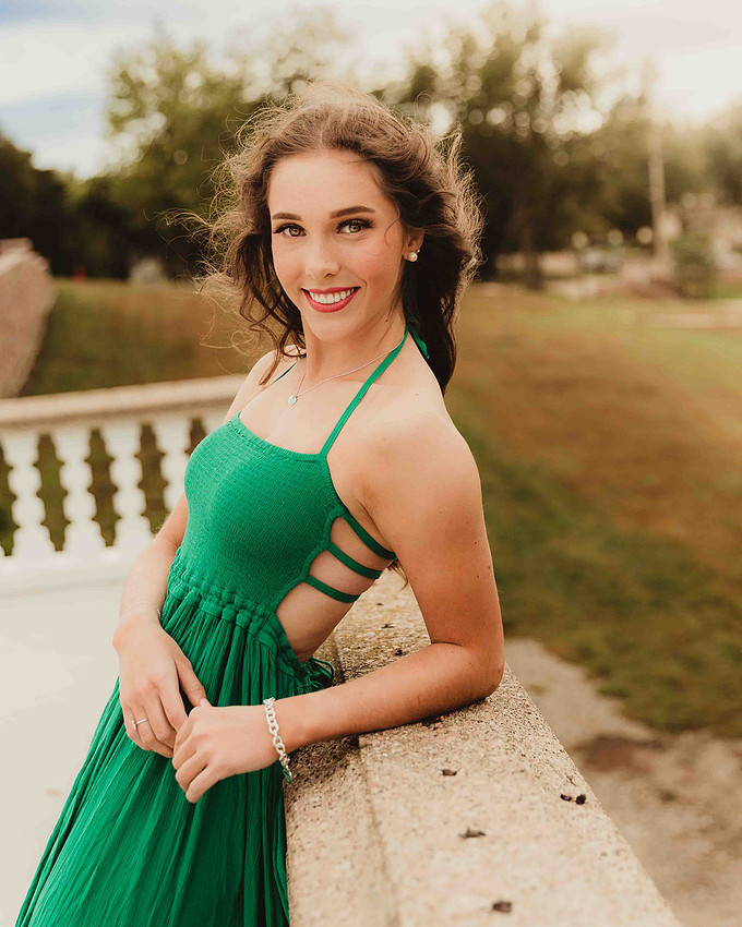 high school senior girl in green maxi dress leaning against garden balcony railing with dark hair blowing in the wind during outdoor senior portrait session in Sioux Falls South Dakota