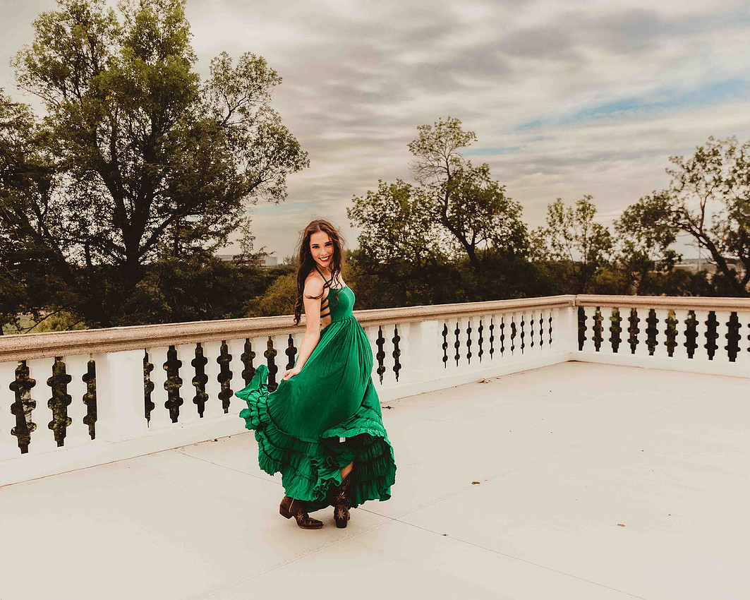 high school senior girl in flowing green maxi dress and cowboy boots dancing on white balcony with gardens and cloudy sky during Sioux Falls senior portrait session