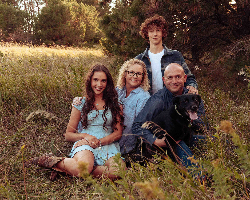 High school senior girl with her family and black Labrador wearing coordinated denim and plaid outfits, standing in a grassy field with pine trees at sunset in South Dakota