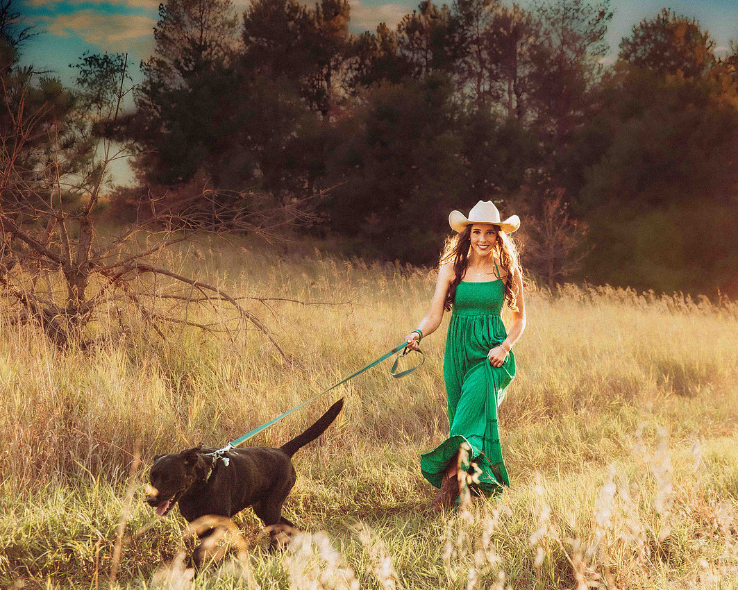 High school senior girl with long flowing hair wearing a cowboy hat and boots walking a black Labrador down a grassy rural road in South Dakota