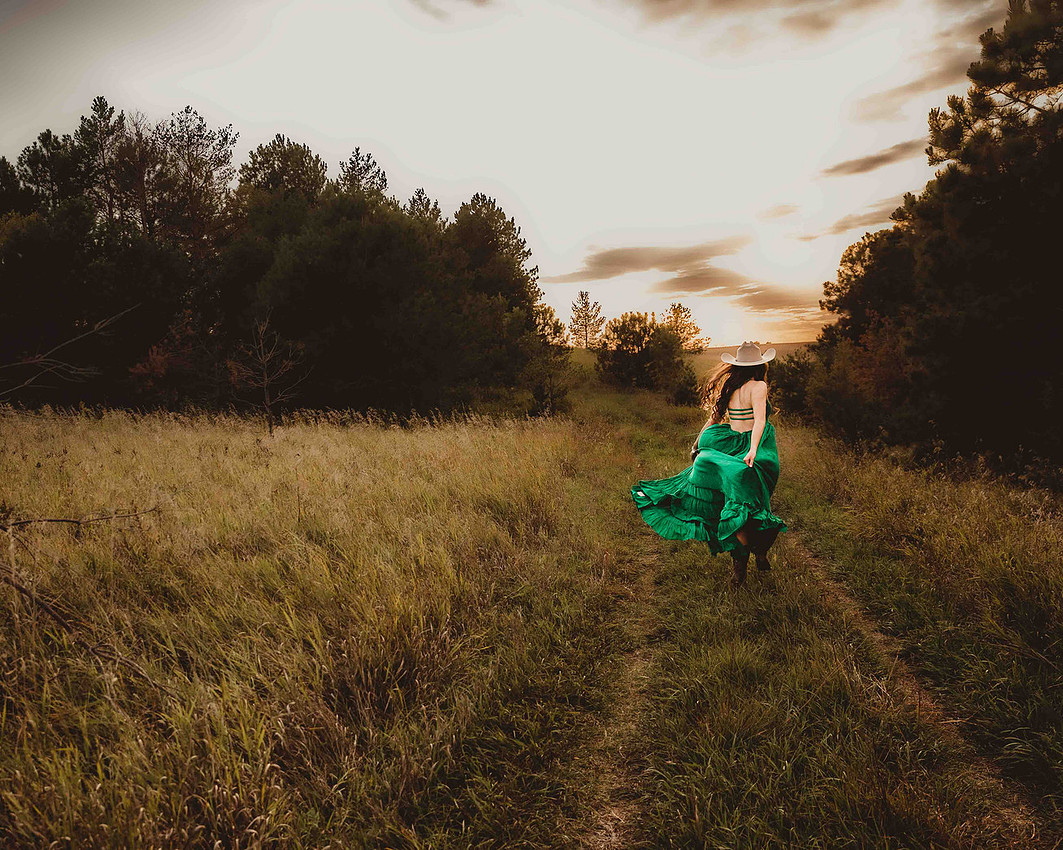 High school senior girl in a flowing green maxi dress, cowboy hat, and boots running down a rural gravel road at sunset in South Dakota