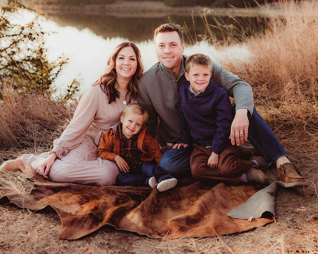 Family of four sitting on a blanket outdoors, smiling at the camera with two young boys during a fall portrait session