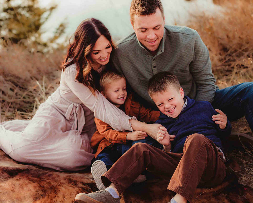 Family of four sitting on a blanket during a fall photo session, parents tickling their two young boys while everyone laughs and smiles with joyful energy