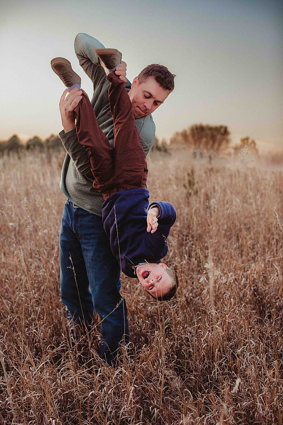 Father holding his young son upside down by the ankles during a family photo session while the boy laughs and giggles, capturing a playful and interactive moment