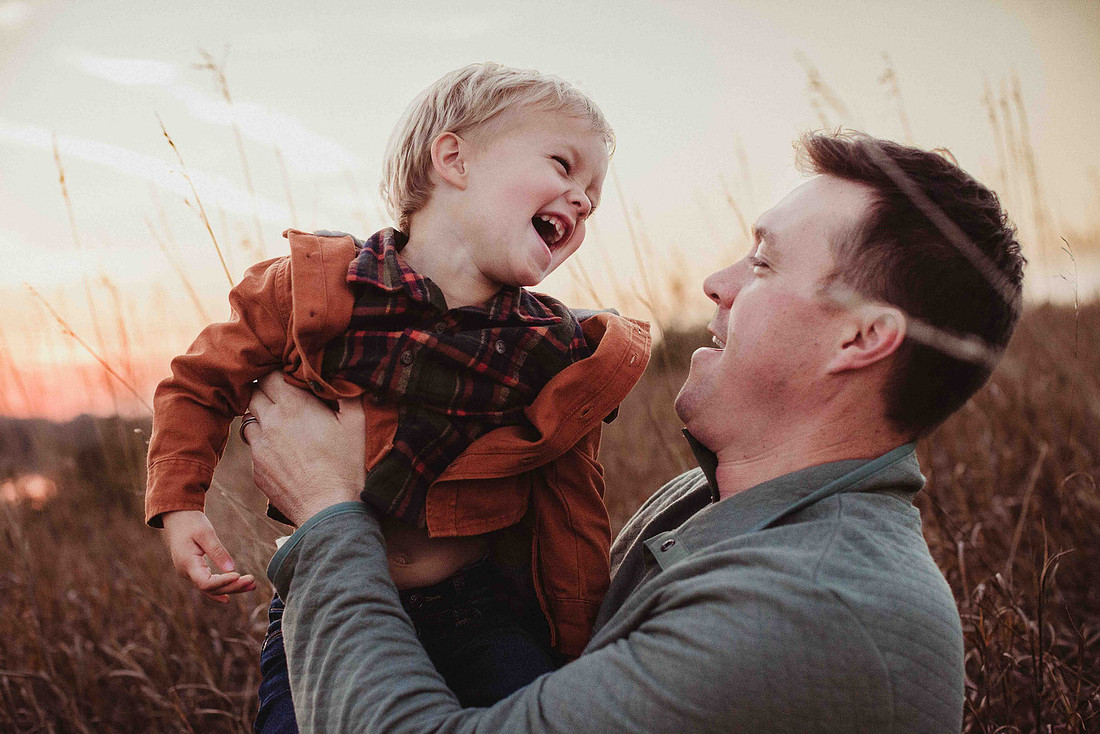 Father holding his young son upside down by the ankles during a family photo session while the boy laughs and giggles, capturing a playful and interactive moment
