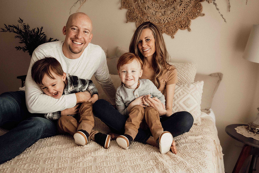 Sioux Falls family of four sitting on a bed, laughing and smiling with parents tickling their young twin boys during a joyful lifestyle photography session