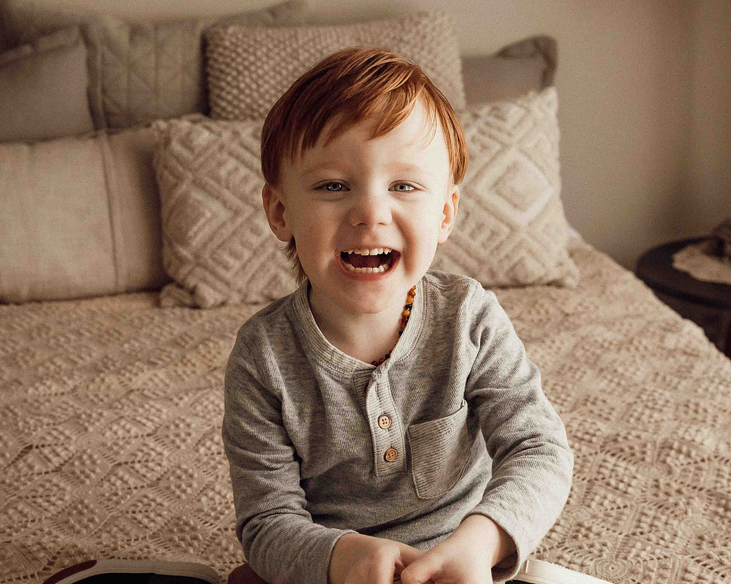 Three-year-old redheaded boy sitting on a neutral cream-colored bed with textured bedding, smiling brightly and full of joy during a lifestyle photo session