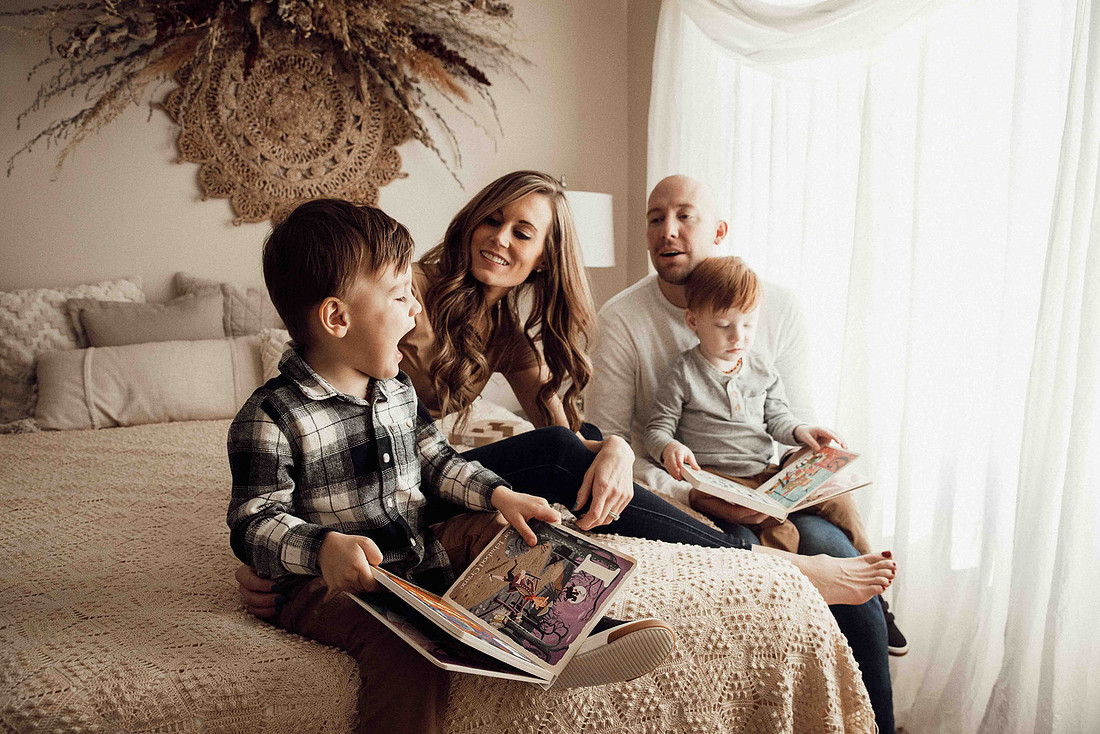 Sioux Falls family photography session with twin toddler boys sitting on a bed by a large window, looking through alphabet books together in soft natural light
