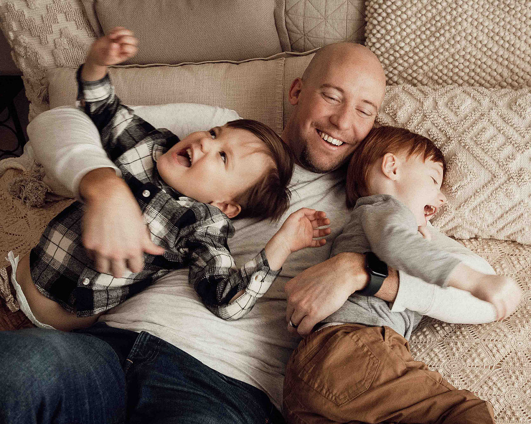 Sioux Falls family session with father lying on his back on a bed, arms around twin toddler boys while tickling them as they laugh and giggle during a playful moment