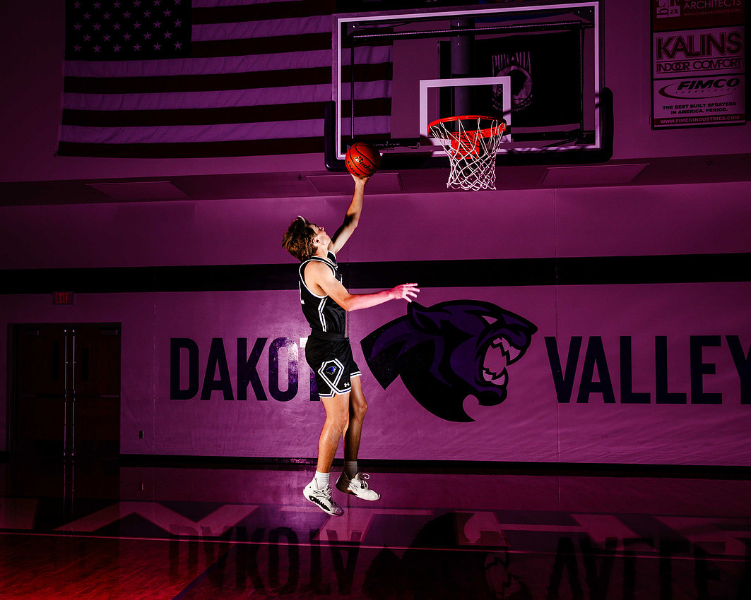 High school senior basketball player captured mid-layup with strobe lighting, featuring a school mascot and American flag in the background with deep purple tones