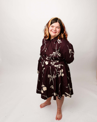 Full-length image of a smiling young person with shoulder-length dark hair and blonde highlights, wearing a maroon floral dress and standing barefoot against a clean, light studio background with a relaxed, confident posture.