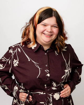 Smiling young person with shoulder-length dark hair and blonde highlights wearing a maroon floral dress, posing confidently with hands at the waist against a clean, light studio background.