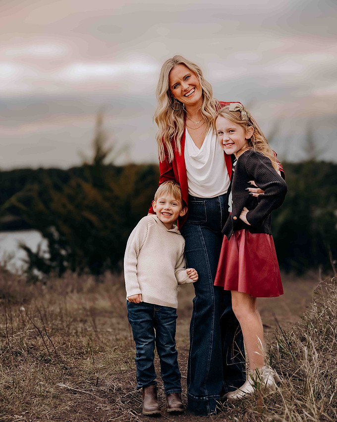 Family smiling outdoors in a fall field near water, wearing coordinated neutral and red outfits during a lifestyle photography session
