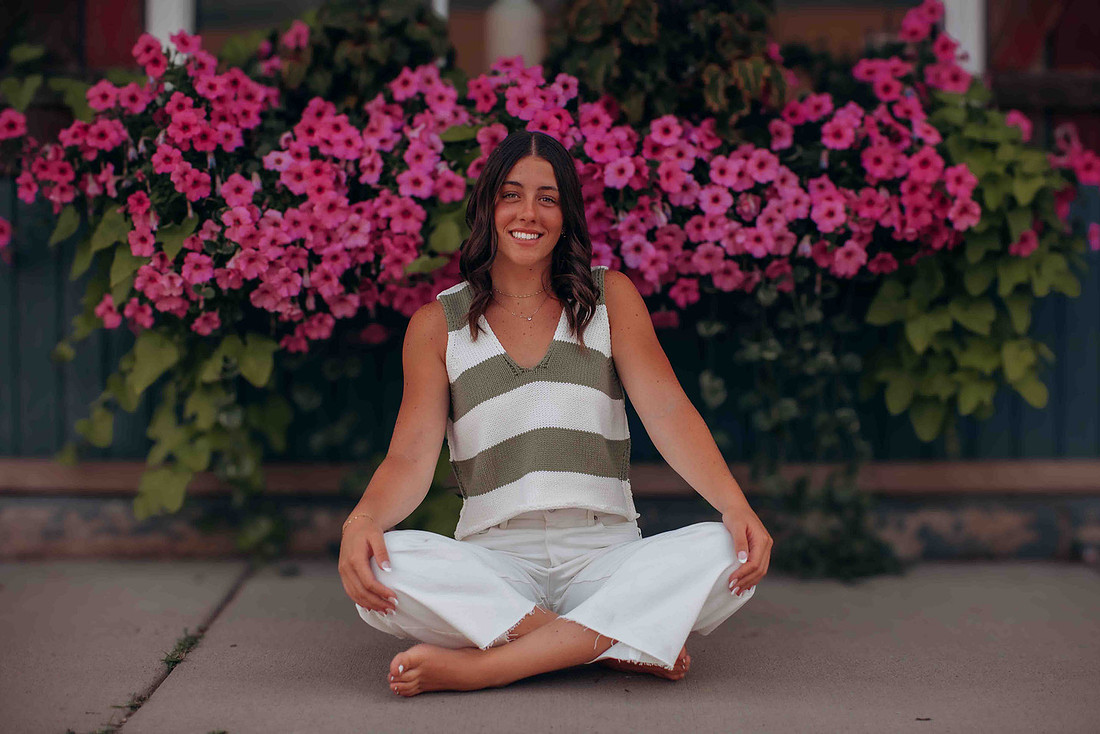 High school senior girl sitting barefoot in front of vibrant pink flowers wearing white jeans and a green striped sleeveless sweater in downtown.