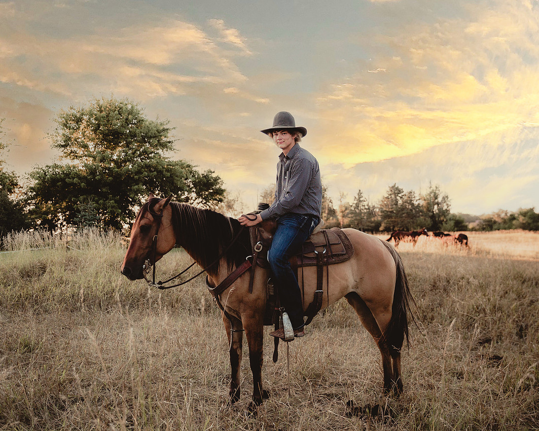 Senior boy wearing a cowboy hat sitting on a saddled sorrel horse in a farm field at sunset during a senior session.
