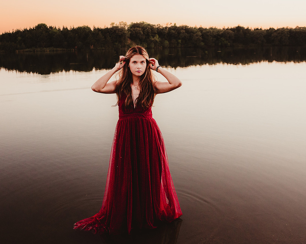 Senior girl in formal gown standing in lake water at sunset with dress flowing in the water during a senior photography session near Sioux Falls South Dakota.