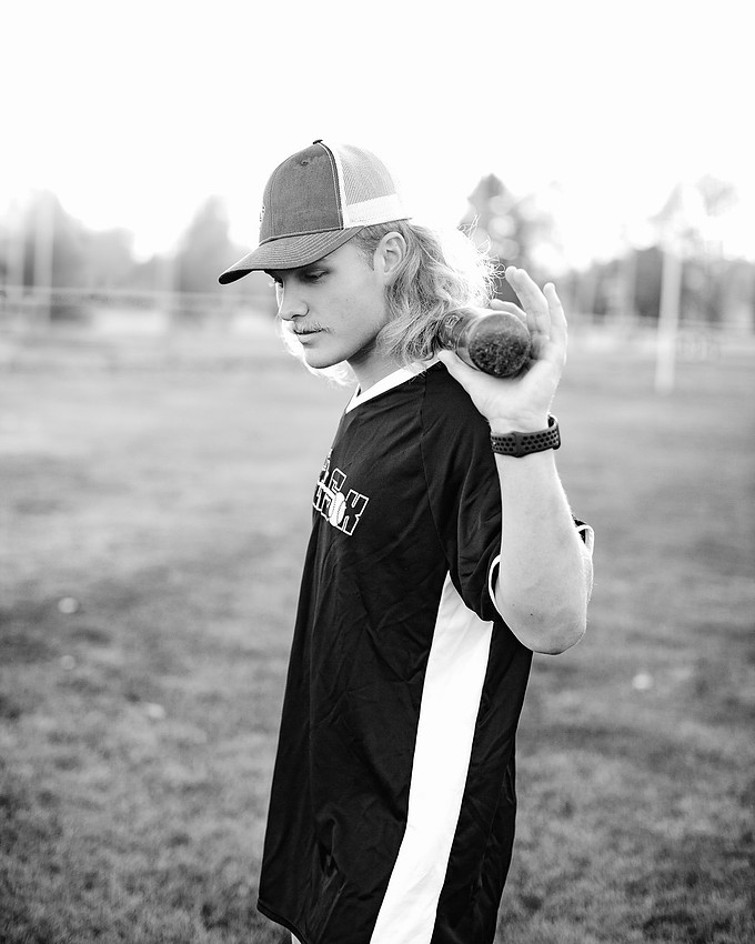 Black and white portrait of a high school senior baseball player in side profile, holding a bat across his shoulders, wearing a cap and jersey with dramatic backlighting