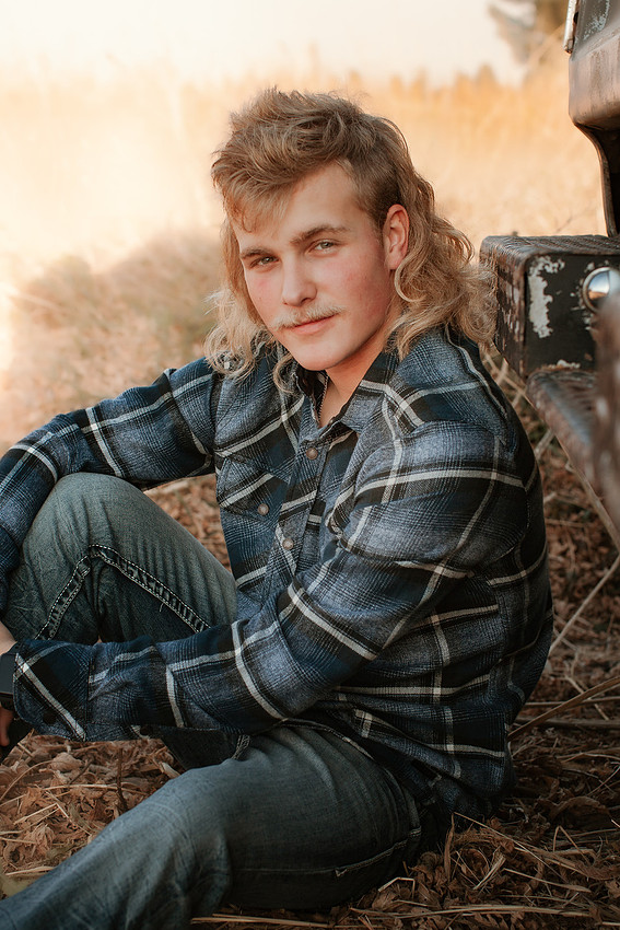 Senior boy with blonde mullet and mustache sitting in front of an old pickup truck in a fall cornfield during golden hour – rustic senior portrait by Sioux Falls senior photographer.